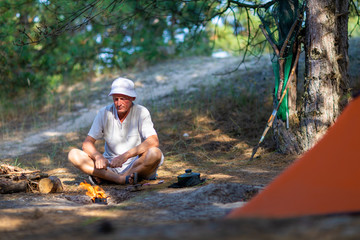 Man looks at a bonfire near a tent in a pine forest