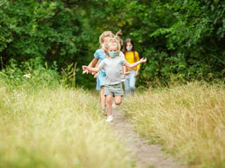 Fototapeta premium Happy little kids wearing protective face mask jumping and running on meadow, forest. Looks happy, cheerful, sincere. Copyspace. Childhood, pandemic concept. Healthcare, coronavirus pandemic.