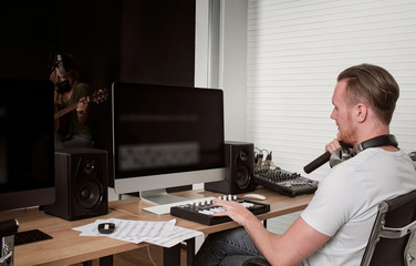 Young Caucasian male close up, music composer playing midi keyboard for arranging song on computer in home studio, female playing guitar and singing on dark background, recording and producer concept.