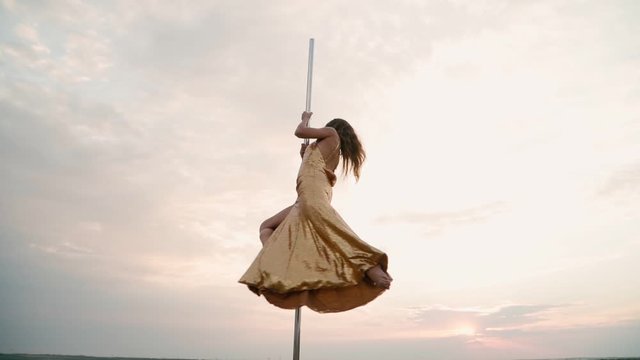 Beautiful athletic woman in a gold dress performs tricks on the pole against the background of sunset in the field. Beauty and body care. Female sports and fitness. Slow motion