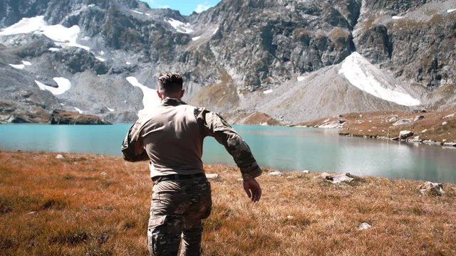 A young soldier in a camouflage uniform runs in the mountains by the lake