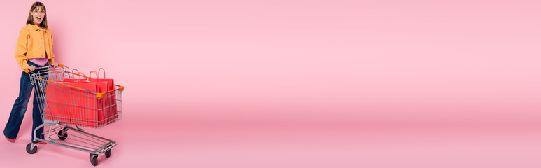 Panoramic shot of excited woman looking at camera near red shopping bags in cart on pink background