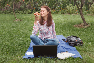 Young woman with a croissant in her hand sitting in the garden on the grass near the laptop. young...