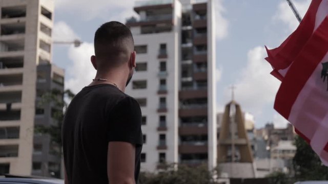 Lebanese adult man wearing face mask standing by red and white Lebanon flags flying in breeze on downtown Beirut city streets after explosion at port, covid-19 pandemic and outbreak, close up behind