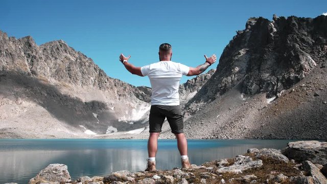 A man traveler in a white T-shirt epic spreads his arms to the sides by a mountain lake