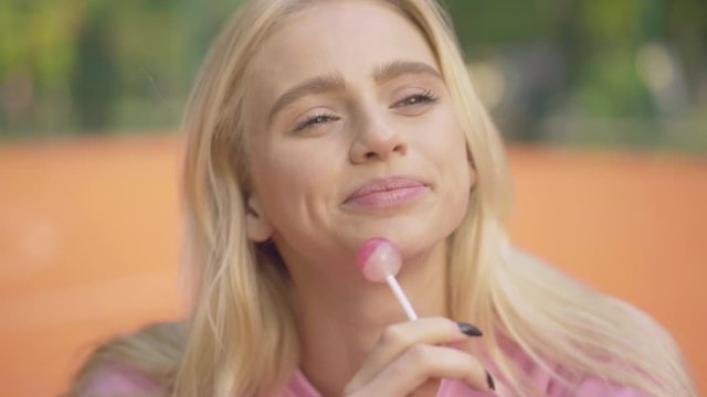 Close-up of blond attractive woman sucking lollipop and laughing. Portrait of beautiful Caucasian joyful lady enjoying sunny summer day on outdoor sports playground.