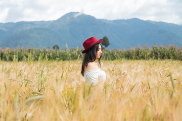 Naklejka na ściany i meble Young woman standing in the middle of a golden wheat field enjoying the sunset - girl with red hat in a field surrounded by mountains - woman alone in the field