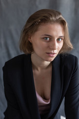 portrait of young caucasian woman with short hair posing in black suit jacket, sitting in front of gray fabric background. closeup shot of pretty girl. short-haired attractive female poses in studio