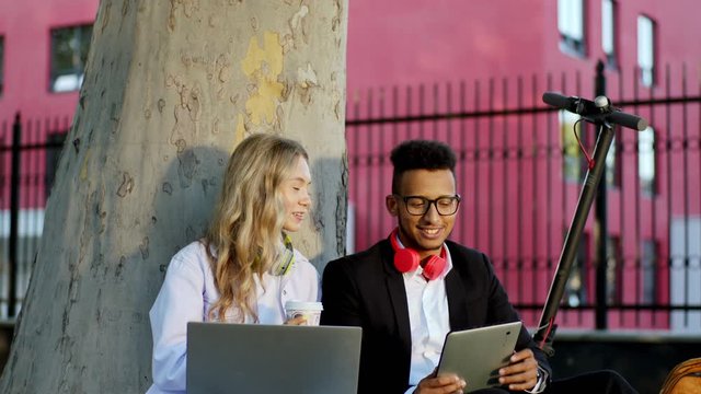 Multiethnic woman and man students have a friendly conversation at the break time they working together at the project using laptop and digital tablet sitting down on the grass beside of a big tree