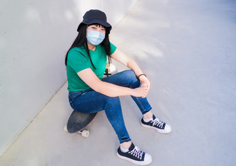 Beautiful young asian woman sitting on a skateboard and wearing protective face mask - Coronavirus lifestyle and health care