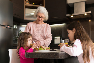 Happy Senior grandmother standing and helping young granddaughter to drink juice at kitchen table, inside. Family, affectionate, reunion, together concept.