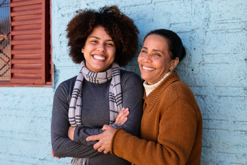 Happy cheerful black adult daughter and mother with broad smile embracing each other outside humble...