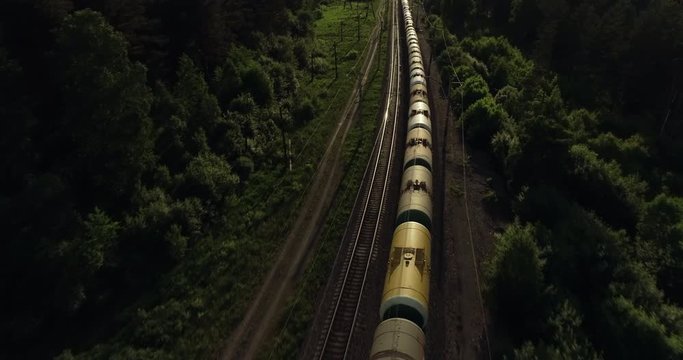 Oil tank and petrol carriages of freight long train carries an electric locomotive by Trans Siberian railways under the rock and near mountain river / Aerial drone wide view at summer sunny day