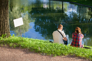 A guy and a girl are sitting on the shore of a pond in a summer park