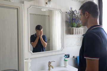 Young man standing in front of the mirror sees himself in desperate reflection with his hands in...