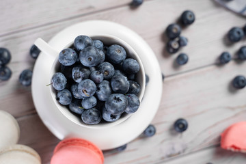 Top view of ceramic cup of ripe blueberries placed on table near sweet macaroons in morning