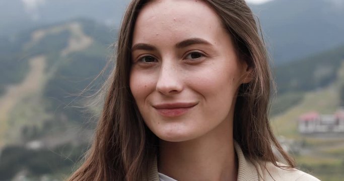 Close up of young woman in blue raincoat spending free time for travelling to mountains. Happy girl with brown hair standing on top of hill after rain.