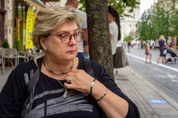 A Mature sad woman in a black dress and glasses looks into the distance.