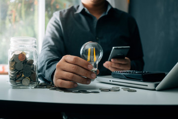 business hand holding lightbulb with using smartphone, laptop computer and money stack in office. idea saving energy and accounting finance concept in morning light