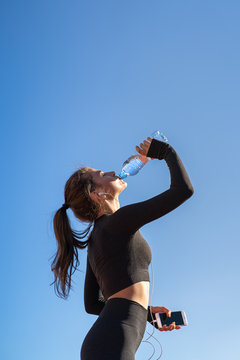 Thirsty young fit woman drinking water from plastic bottle, resting after jogging, holding mobile smartphone in her hand, listens to music. Blue sky with copy space. Hot day. 