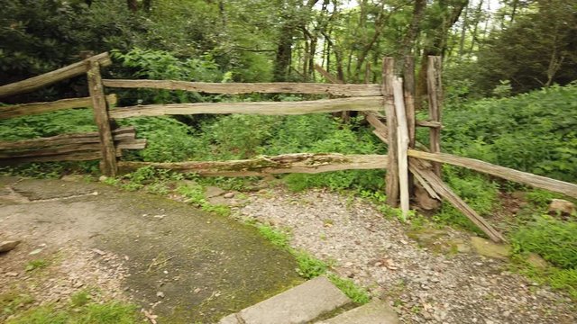 Scenic old rustic damaged split rail fence by spring house farm building in rural countryside farmland by green grass, vegetation and tree forest on sunny day, handheld pan left