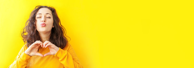 Love heart shape peace. Beauty portrait young happy woman showing heart sign with hands on yellow background isolated. European girl. Positive human emotion facial expression body language Banner