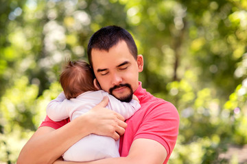 Father with closed eyes tenderly embricing infant daughter in pink jumpers outdoors in warm sunny or early autumn day. Baby is hugging dad too. Parental love, care and tenderness concept