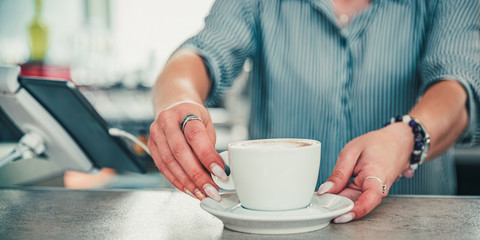 Fototapeta na wymiar Closeup female barista hands holding cup of aromatic latte or cappuccino coffee in bar or cafe