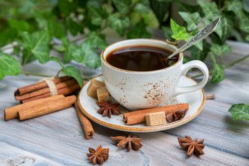 White cup of coffee with cinnamon and aniseed stars on a light wooden background with green ivy. Selective focus