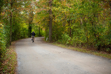 A senior man riding his bicycle down a gravel trail beneath colorful fall leaves in Belmont Lake State Park, Long Island, New York