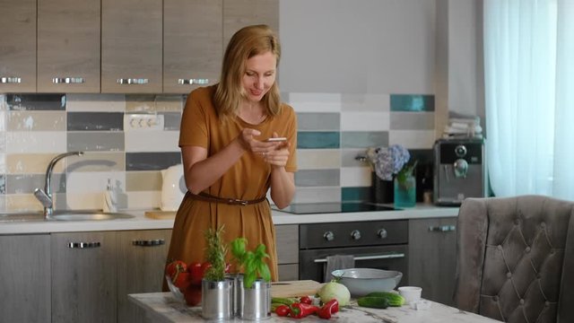 Young caucasian woman is taking pictures of food while standing by table in home kitchen rbbro. Beautiful female makes photo of fresh vegetables on desk and smiles, holding smartphone in hand in light