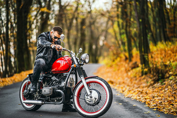 Obraz na płótnie Canvas Bearded brutal man in sunglasses and leather jacket sitting on a motorcycle on the road in the forest