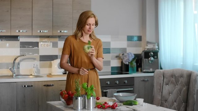 Young woman talking about nutrition and showing vegetables while standing at table in home rbbro. Beautiful lady speaking with smile and holding organic food in hands at desk in light interior