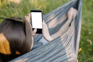 Young woman chilling in hammock and holding phone with empty screen. Girl with phone chatting with friends or making order online, while relaxing in hammock. Phone mock up