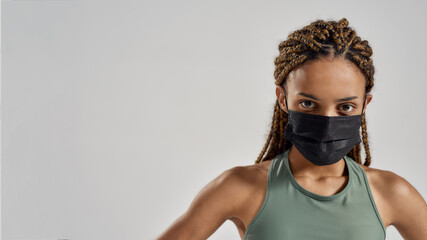 Coronavirus and sport. Confident sportive mixed race woman wearing black medical protective face mask looking at camera while standing isolated over grey background