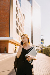 Young busy woman wearing business clothes, standing on modern buildings background, talking on the smartphone.