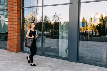 Young modern woman wearing business clothes, walking on glass wall background, holding documents.