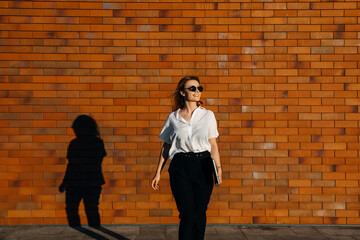 Young stylish woman wearing white shirt and black pants, walking on the street, on a brick wall background, with a laptop, smiling.