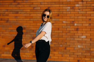 Young modern woman wearing sunglasses, white shirt and black pants, walking on a brick wall background outdoors, smiling.