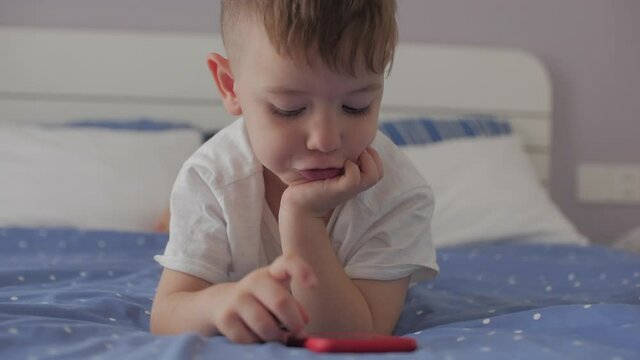 Cute little boy preschool child using a phone or digital tablet technology device looks telephone,plays in the phone sitting rest lying on sofa alone.Children tech addiction concept play game at home.