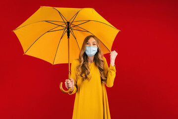 Happy young woman, in a yellow dress and a medical protective mask on her face, with an umbrella in her hands, shows a winning gesture, on a red background