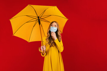 Pensive young woman in a yellow dress and a medical protective mask on her face, with an umbrella in her hands, looks to the side, on a red background