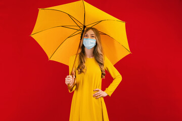Beautiful young woman in a yellow dress, in a medical mask on her face, with an umbrella, on a red background