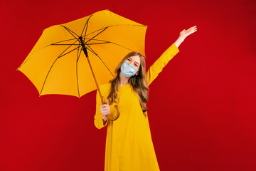 Happy girl in a yellow dress and a medical protective mask on her face is having fun, with an umbrella in her hands, on a red background