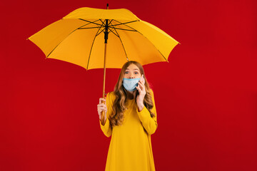 shocked girl in a surgical protective mask on her face, in a yellow dress and with a yellow umbrella in her hands looks surprised and talks on a mobile phone on an isolated red background