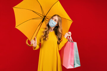 Pensive young woman in a medical mask, with shopping bags and an umbrella, on a red background