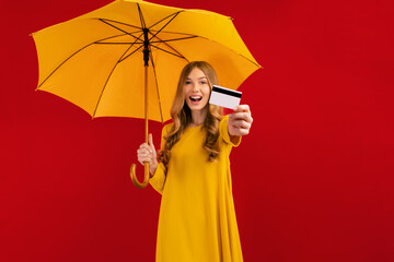Shocked happy young woman, with umbrella and credit card in hand, on a red background