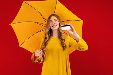 Happy beautiful young woman with umbrella and credit card in hand, on a red background