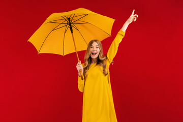 Excited young woman with a yellow umbrella having fun and showing a winning gesture on a red background
