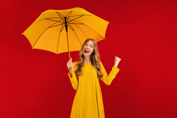 Excited young woman with a yellow umbrella having fun and giving a winning gesture on a red background